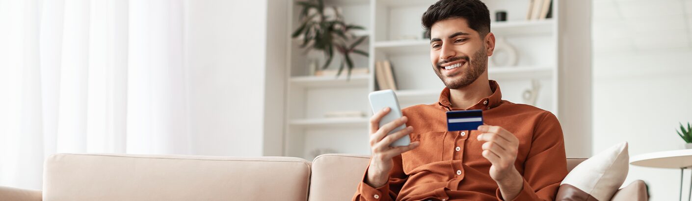 Happy man sitting on the couch with his cell phone in one hand and a credit card in the other.