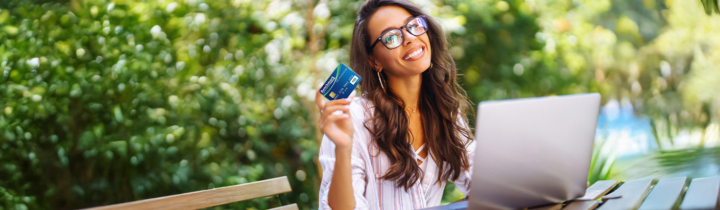 Close up of an adult female working on a laptop outside while holding a BFCU credit card in the air.