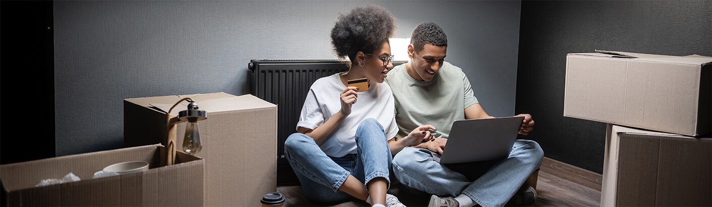 Excited couple sitting on the floor of a new living space while viewing a laptop computer.