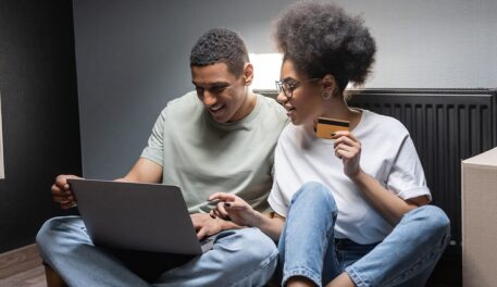 Excited couple sitting on the floor of a new living space while viewing a laptop computer.